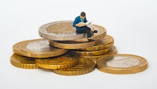 A small doll reading a book whilst sitting on top of a pile of coins